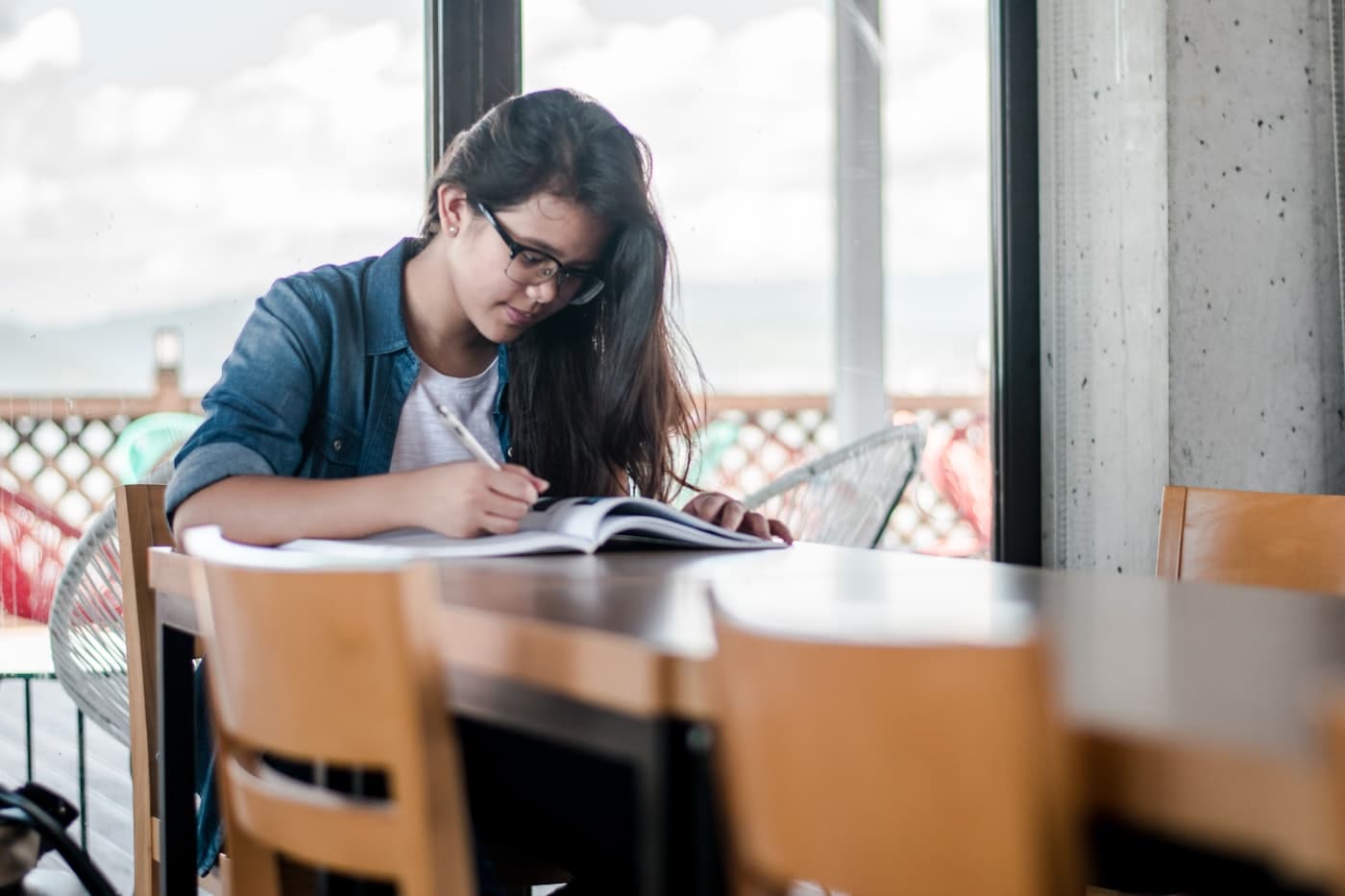 A student writing in an open notebook at a table beside a large window
