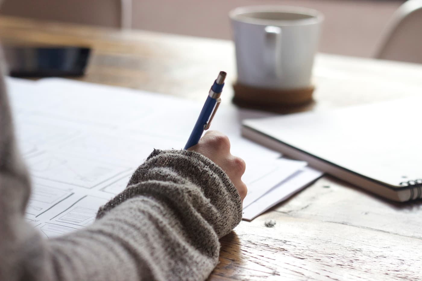 A close-up of a hand holding a pen over handwritten notes, beside a mug and a notebook